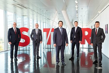 Photo of the five members of the STRABAG SE Management Board in front of a glass front in the STRABAG building