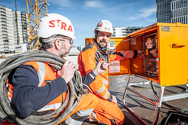 Foto von zwei STRABAG Bauarbeiter die zusammen an einem Stromkasten auf einer Baustelle arbeiten. 