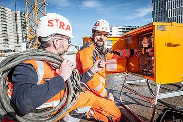 Photo of two STRABAG construction workers working together on an electricity box on a building site. 