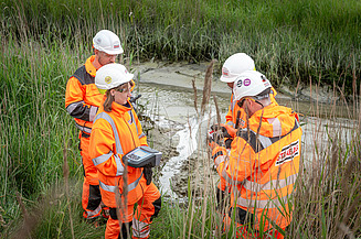 The inspection by four STRABAG employees of a body of water in a green landscape Photo shows four STRABAG employees standing by a body of water in a green landscape and checking it